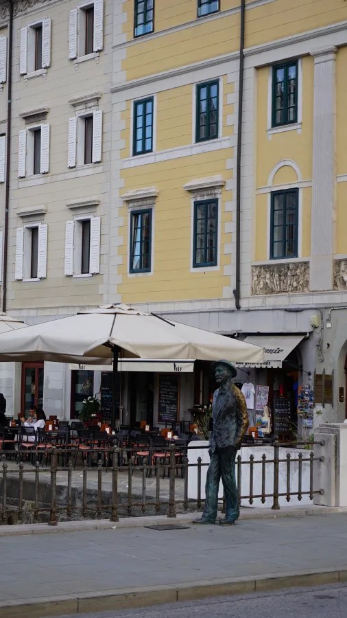 Bronzeskulptur eines Mannes in einem Stadtbild mit farbigen Gebäuden und Cafés.