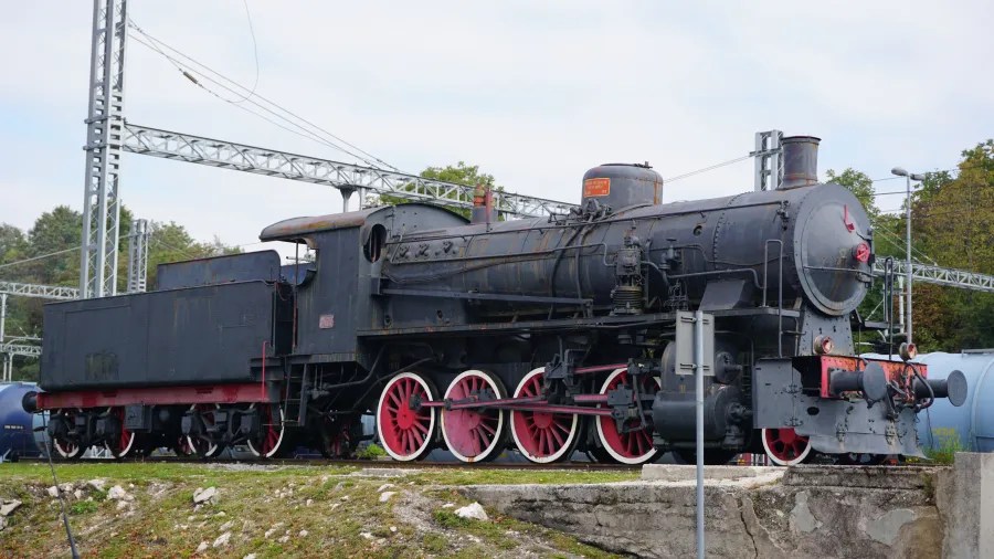 Eine historische Dampflokomotive mit roten Rädern, abgestellte auf einem Gleis, umgeben von technischen Anlagen und Bäumen im Hintergrund.