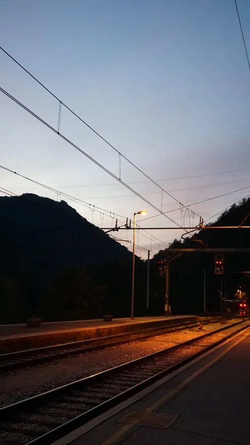 Bahnhof mit Gleisen und einem beleuchteten Signal bei Dämmerung, umgeben von Bergen.