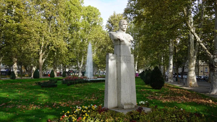 Eine Wiese mit einer Statue eines Mannes in einem Stadtpark, umgeben von Bäumen und Blumenbeeten.