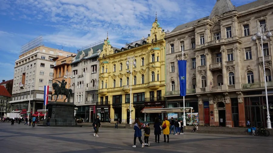 Stadtplatz mit historischen Gebäuden, darunter ein gelbes Gebäude und eine Statue eines Reiters, unter bewölktem Himmel.