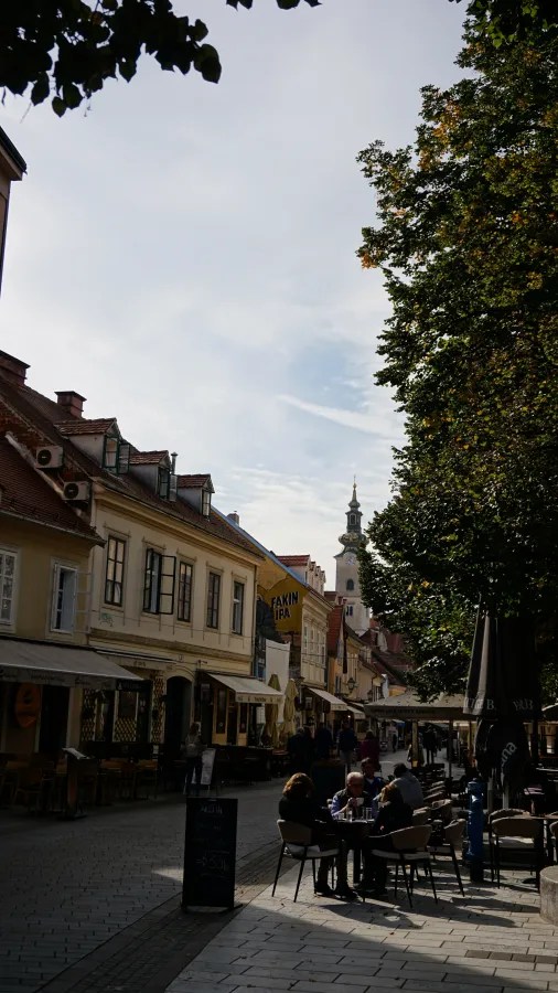 Blick auf eine lebhafte Straße mit Cafés und Restaurants, umgeben von historischen Gebäuden, und einem Kirchturm im Hintergrund unter blauem Himmel.