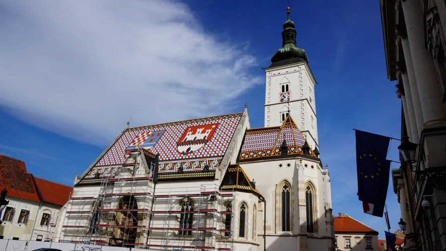 Das Bild zeigt eine beeindruckende Kirche mit einem bunten Dachmuster und einem hohen Glockenturm. Vor der Kirche befinden sich Baugerüste und im Hintergrund sind blaue Himmel und europäische Flaggen zu sehen.