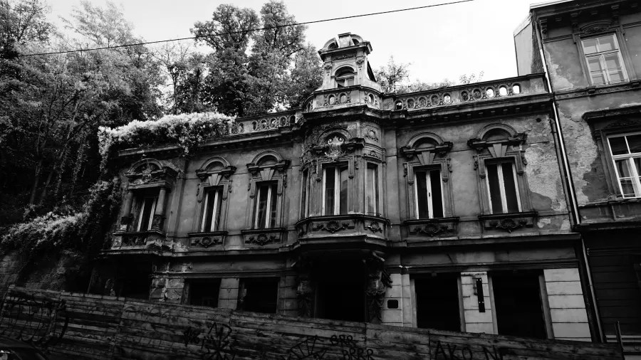 Verfallenes historisches Gebäude mit bemoosten Wänden und kunstvollem Balkon, umgeben von Bäumen.