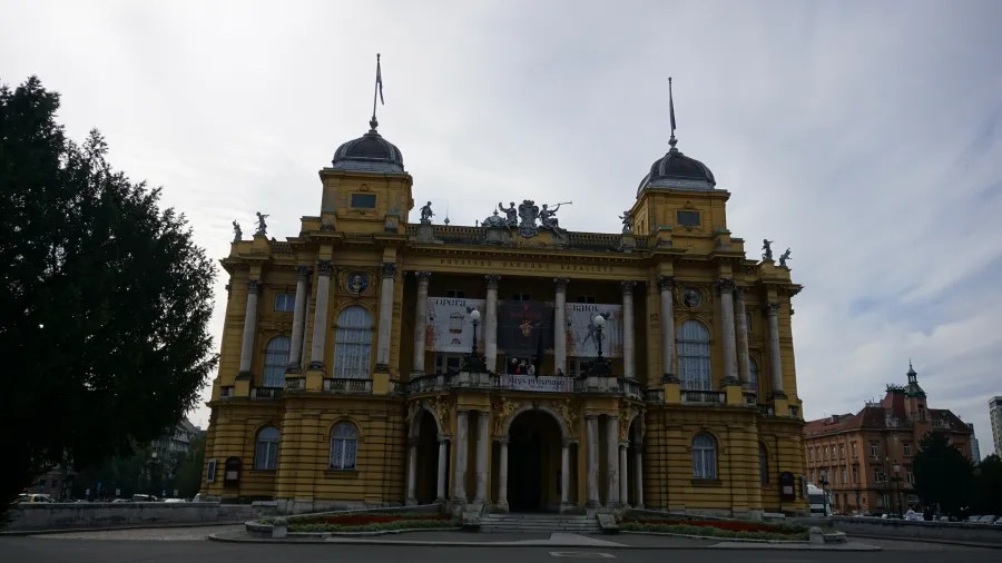 Historisches gelbes Gebäude mit Säulen und Verzierungen, das Theater zeigt, unter einem bewölkten Himmel.