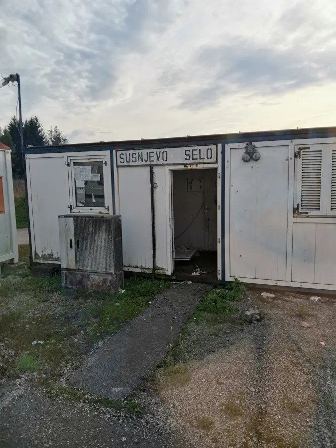 Ein verlassener Container mit dem Schriftzug 'Susnjevo Selo' an der Vorderseite, umgeben von Gras und Schotterwegen.