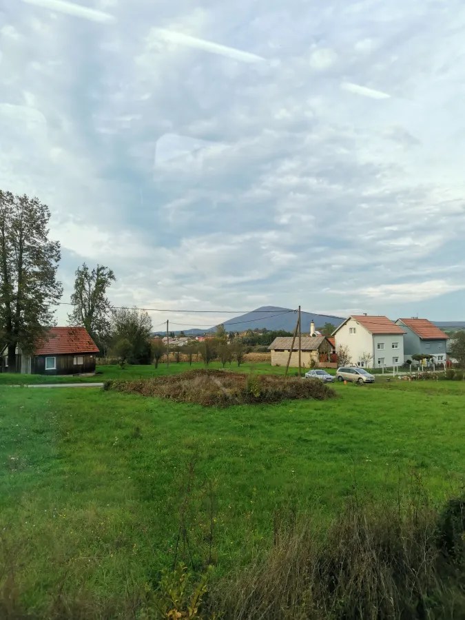 Landschaft mit Wiesen, Häusern und einem Hügel im Hintergrund unter einem bewölkten Himmel.