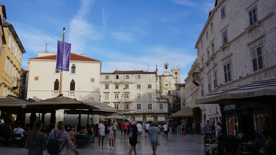 People walking through a historic stone square with outdoor cafes, a clock tower, and Palača Judita.
