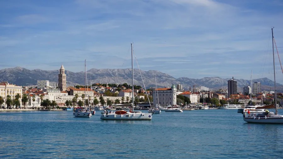 Sailboats in a harbor with a historic coastal city and rugged mountains in the background
