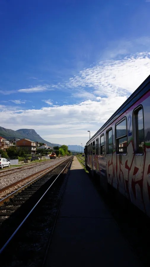 A graffiti-covered train at a platform beside tracks with mountains in the background.