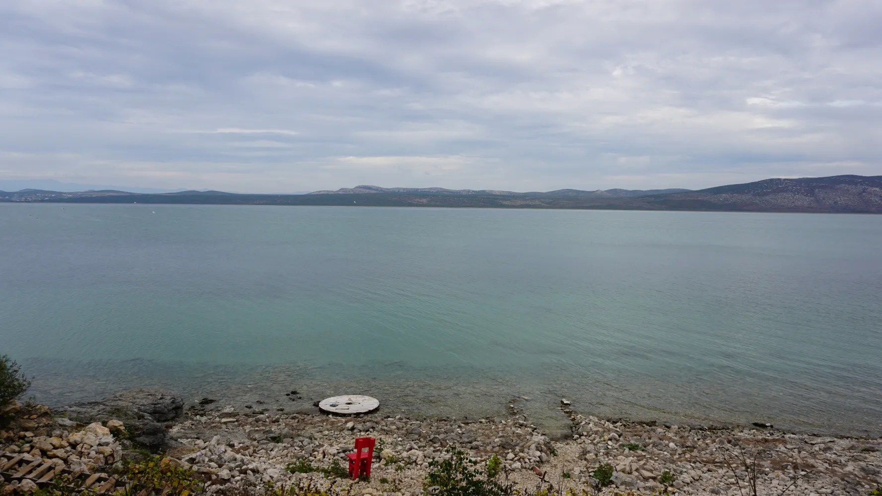 Calm turquoise lake with a rocky shore, a small red chair, and distant rolling hills.