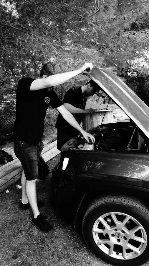 Two people inspecting a car's engine with the hood open in a wooded area