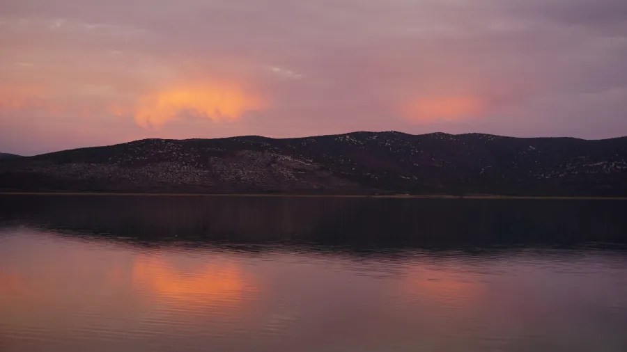 A calm lake reflecting a pink and orange sunset sky over dark hills