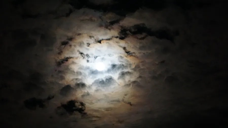 Bright full moon shining through dark, textured clouds in the night sky.