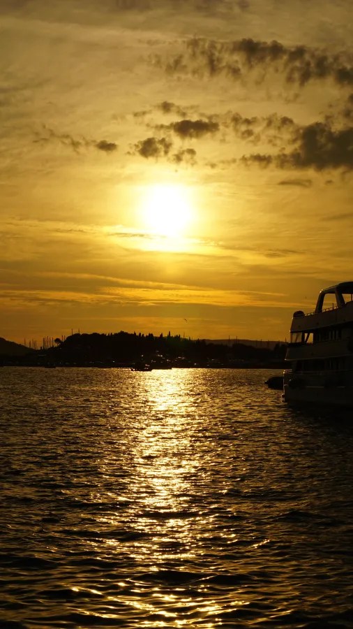 20221009_Kroatien_Split (5) Golden sunset reflecting on water with a silhouetted boat and coastline in the distance.