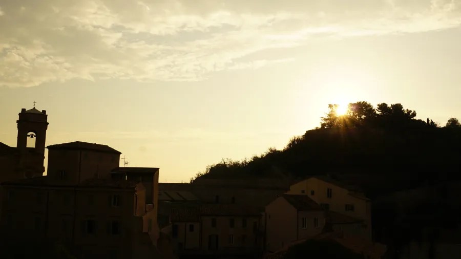 Sun setting behind a wooded hill overlooking a silhouetted European town with a church tower.
