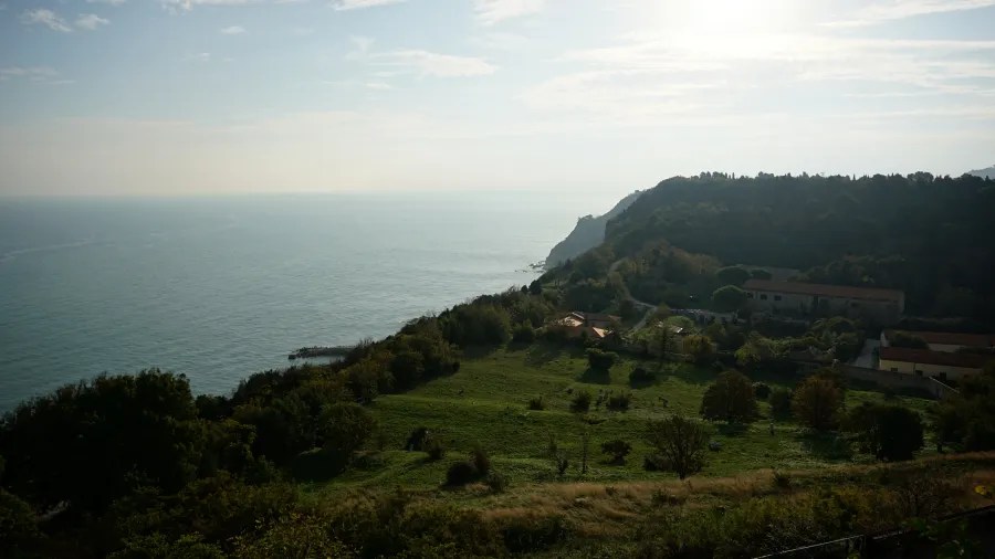 Lush green hills with scattered buildings overlooking a vast blue sea under a bright sky.