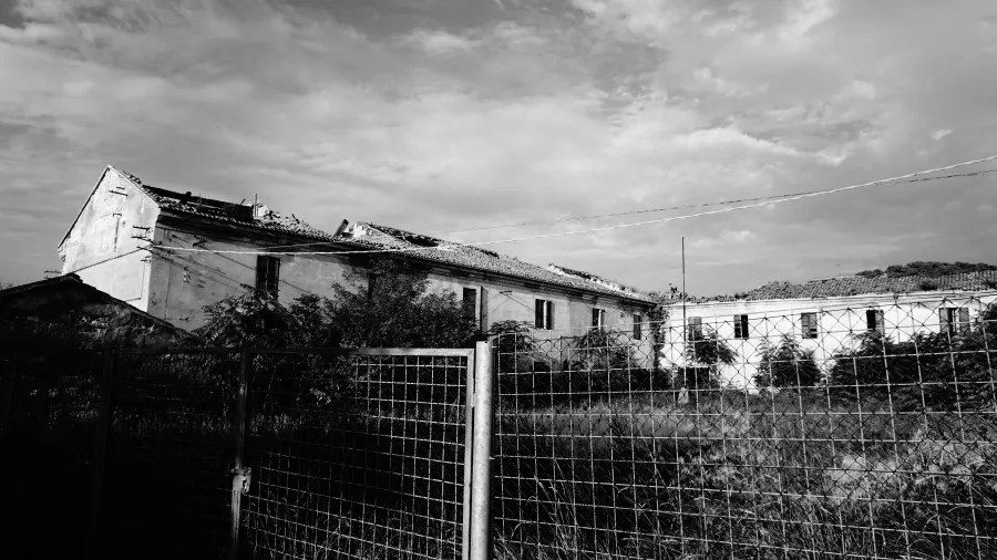 A dilapidated abandoned building with a collapsed roof behind a chain-link fence.