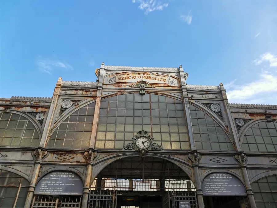 The iron and glass facade of the Mercato Pubblico in Ancona, dated 1926, with a central clock. Left sign: COMUNE DI ANCONA MERCATO DELLE ERBE PRODOTTI COMMERCIALI PIANO SUPERIORE: ALIMENTARI PANE SALUMI, CARNI (BOVINE SUINE OVINE CONGELATE), POLLAME, FORMAGGI LATTE LATTICINI, FRUTTA ORTAGGI DROGHERIA, PESCE FRESCO (DI FRONTE INGRESSO V. MAGENTA). Right sign: COMUNE DI ANCONA MERCATO DELLE ERBE PRODOTTI COMMERCIALI PIANO TERRA: ALIMENTARI GASTRONOMICI VARI, CARNE (BOVINE SUINE EQUINE OVINE), POLLAME, SALUMI VINI E OLIO, FORMAGGI LATTE LATTICINI, ORTOFRUTTICOLI FIORI DROGHERIA, ATTIVITA' DI BAR.
