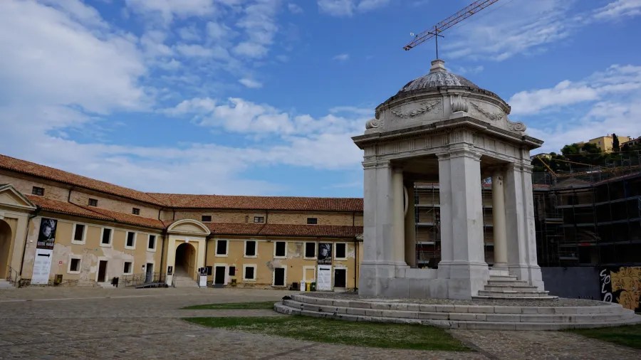 Large white stone domed monument in a courtyard with a long yellow building