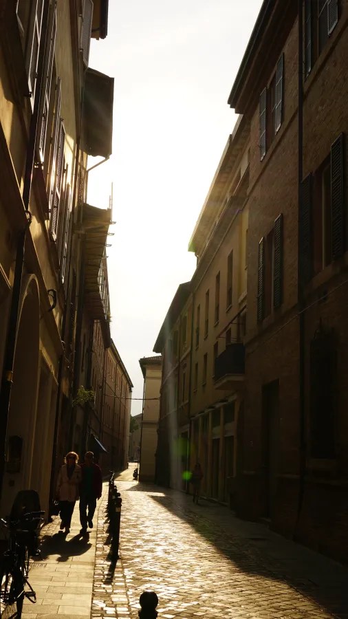 Sunlight streaming down a narrow cobblestone street lined with historic buildings and people walking.