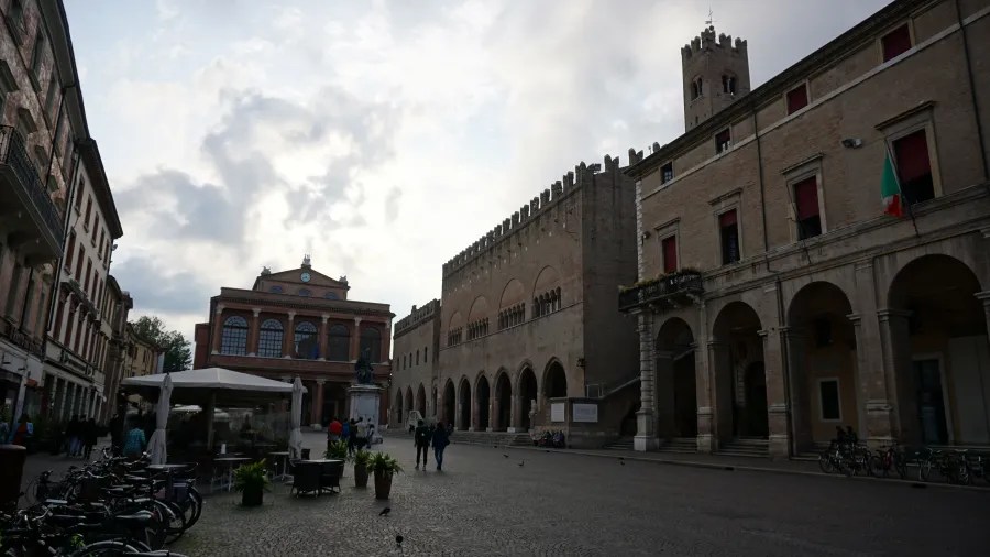 Historic European city square with medieval stone buildings and people walking under a cloudy sky