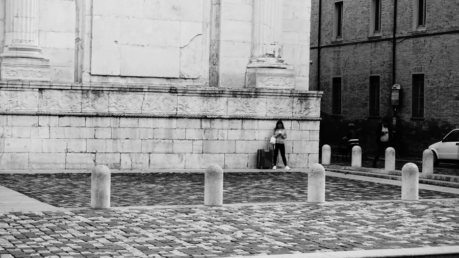A person with a suitcase stands against a large stone building, looking at their phone.