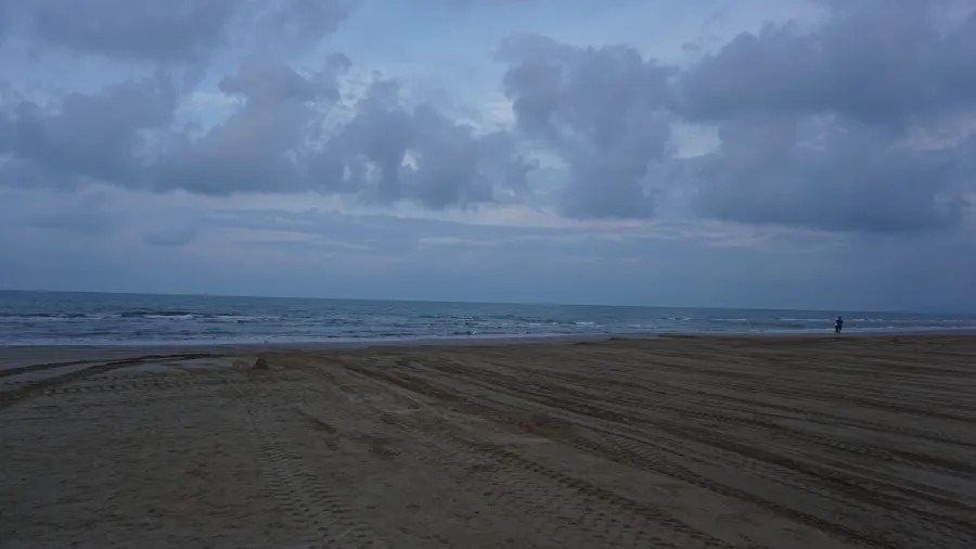 A wide sandy beach with tire tracks under a dark, cloudy sky at dusk