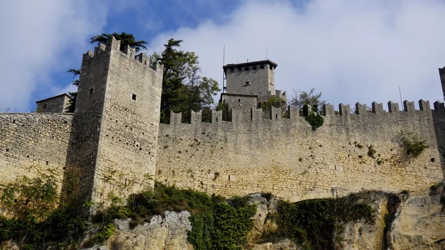 Crenelated stone walls and towers of a medieval fortress on a rocky cliff.