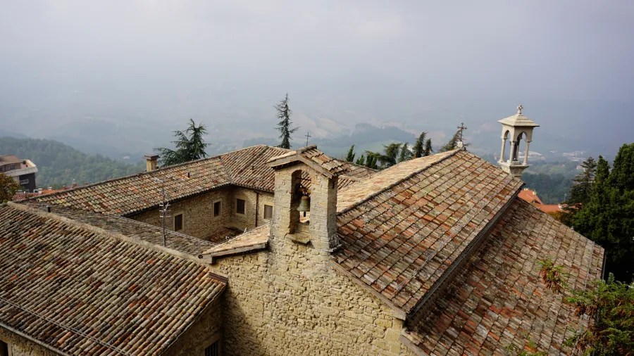 Stone building with terracotta tiled roofs and a bell tower overlooking a misty valley