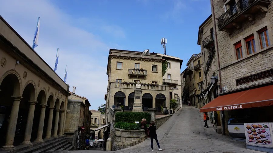 A stone plaza with historical buildings, a statue, and Bar Centrale under an orange awning.