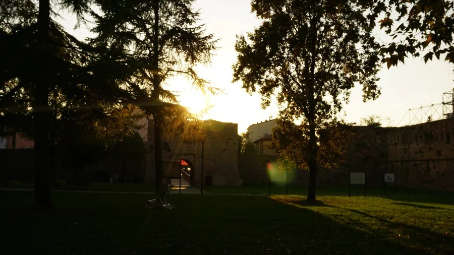 The sun setting behind silhouetted trees in a park with an ancient stone wall.