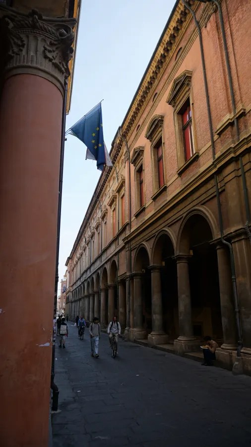Narrow street lined with historic brick buildings, arched porticos, and people walking and cycling.