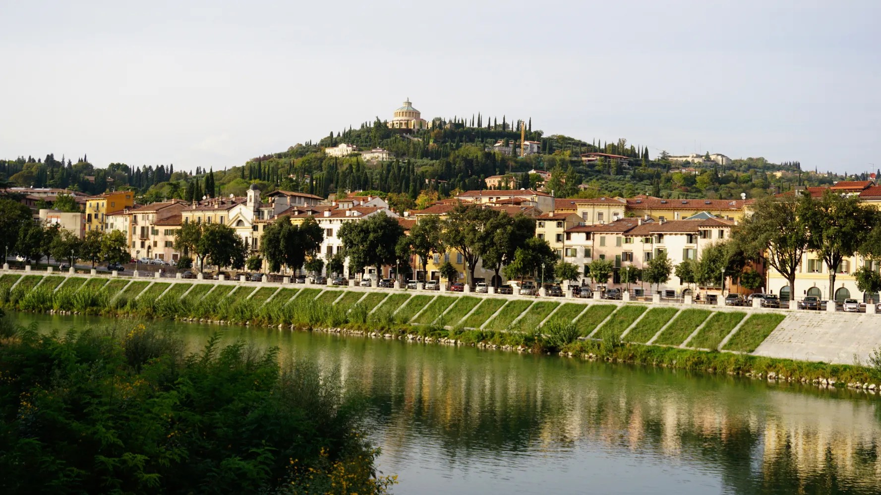 Riverfront buildings in a city with a domed monument on a forested hill backdrop.