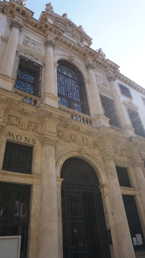 Classical stone building facade with columns, sculptures, and inscriptions reading MONS PIETATIS and ΨΥΧΗΣ ΙΑΤΡΕΙΟΝ.