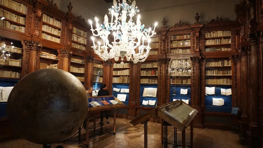 Historic library room with ornate wooden bookshelves, a large globe, and a central glass chandelier.