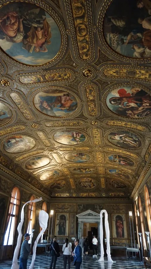Grand hall featuring an intricate gilded ceiling with multiple paintings and modern fabric installations.