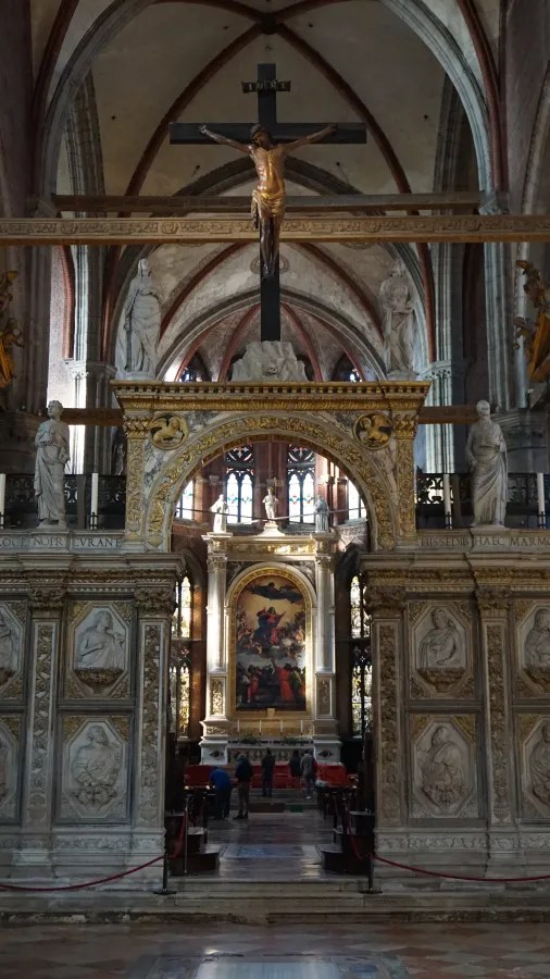 Large hanging crucifix and ornate marble screen framing a view of Titian's Assumption painting