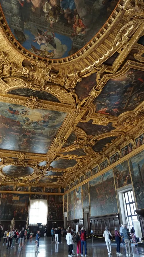 Ornate room with a gilded, painted ceiling and large historical paintings lining the walls.