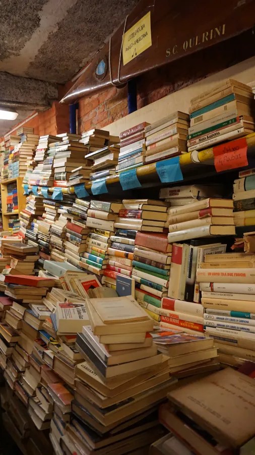 Densely packed stacks of books on shelves in a bookstore, with a boat hull above labeled LETTERATURA ANGLO-AMERICANA and S.C. QUERINI.