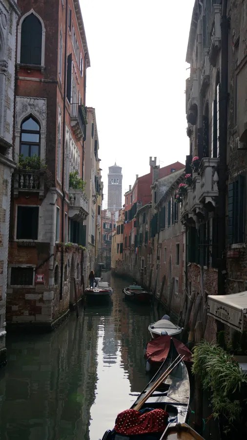 Narrow Venice canal lined with historic buildings and boats leading to a distant bell tower