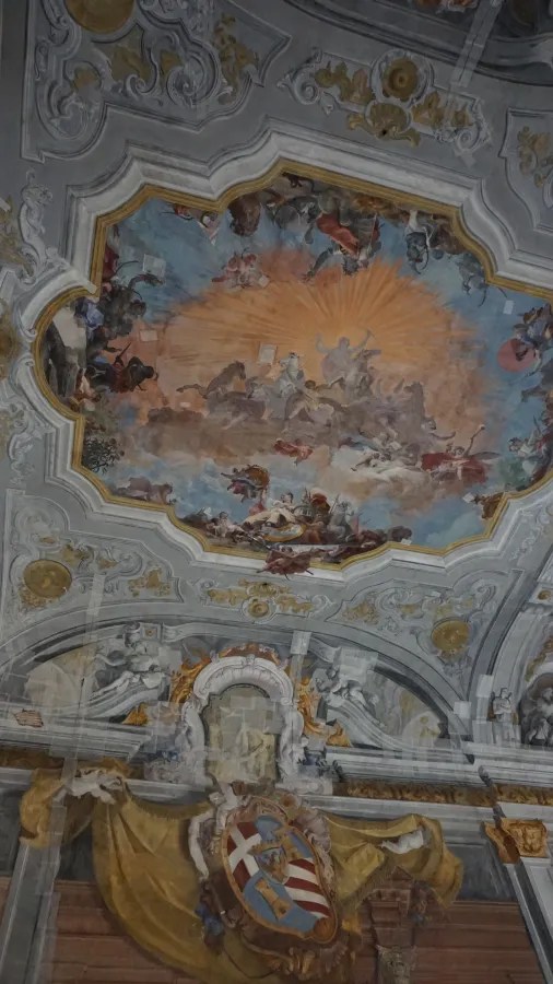 Ornate baroque ceiling featuring a central fresco of mythological figures and a large coat of arms.