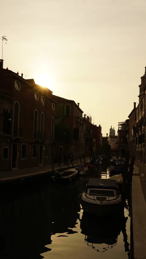 Venice canal at sunset with silhouetted buildings and moored boats reflecting in the water