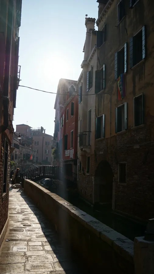 Sunlit narrow canal in Venice with colorful historic buildings and a small bridge.