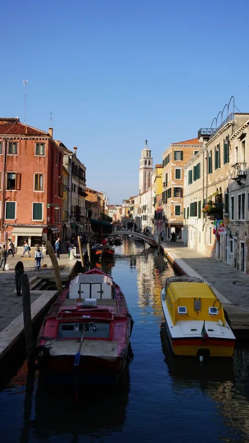 A narrow Venice canal lined with historic buildings, docked boats, and a distant bell tower.