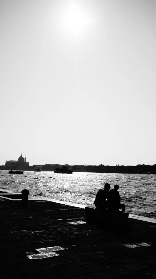 Silhouetted figures sitting on a dock overlooking a sparkling canal in Venice.