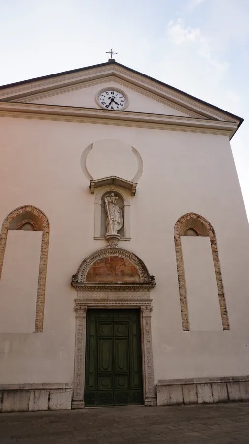 20221015_2_Italien_Udine (14) A white church facade featuring a clock, a central saint statue, and a green door.