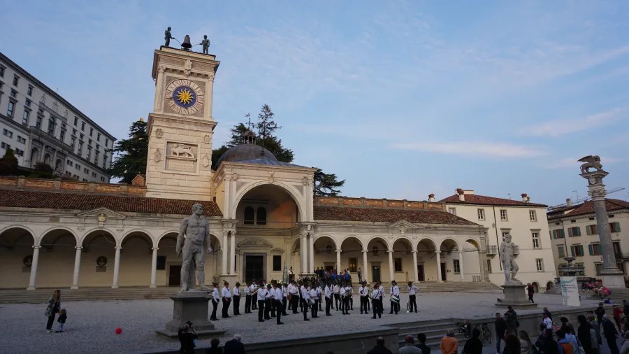 20221015_2_Italien_Udine (38) A brass band performs in a historic square with a prominent clock tower and statues.