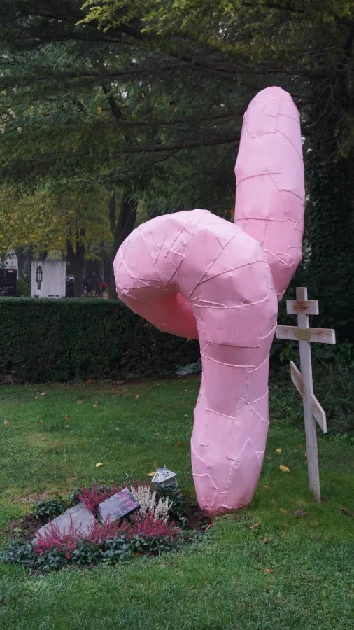 Large pink abstract sculpture standing beside a grave with a wooden cross in a cemetery.