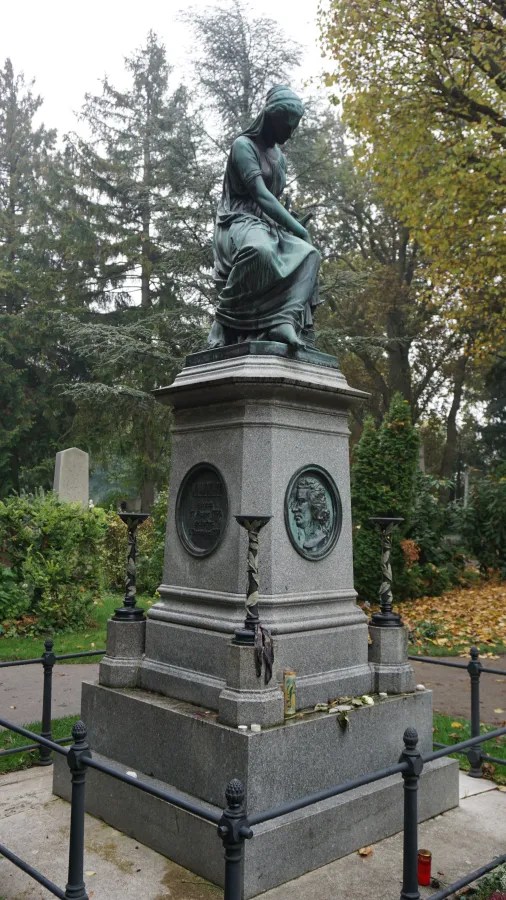 Bronze statue of a seated woman atop a large stone pedestal in a wooded cemetery